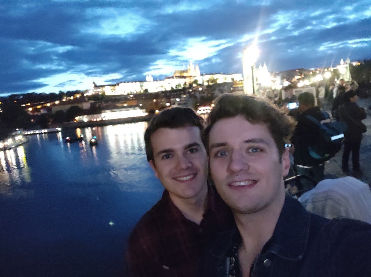 Ale and Harry on a bridge in Prague at dusk, the lit castle and the river behind them.